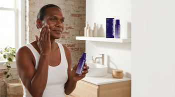 Person with deep brown skin applying an exosome leave-on treatment in a bright minimal bathroom, morning light