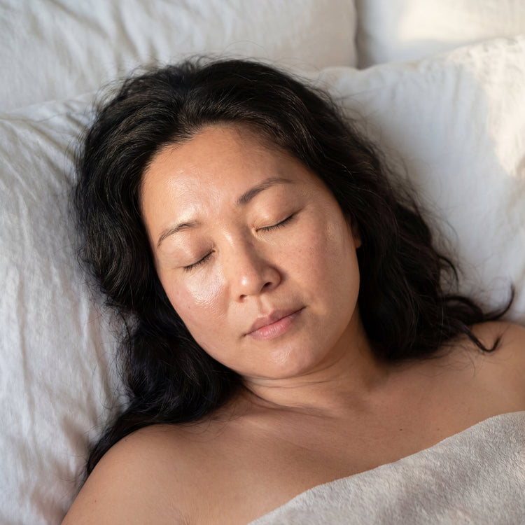 34-year-old East Asian Australian woman with medium skin tone and curvy figure resting peacefully on white bedding, eyes closed, skin appearing healthy and dewy in warm bedroom lighting