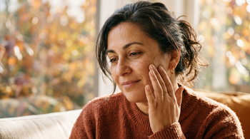 45-year-old Southern European Australian woman with olive skin gently touching her cheek, examining subtle pigmentation in warm autumn light