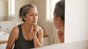 47-year-old Pacific Islander Australian woman with very fair skin examining her face in a bathroom mirror, fine lines visible around her eyes, soft natural lighting